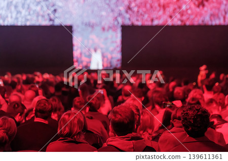 Audience in red lit theater watching live performance on large stage, crowded back view at event Audience in red lit theater watching live performance on large stage, crowded back view at event 139611361