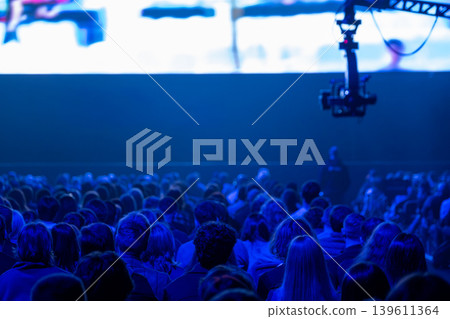 Audience watching conference presentation on large screen, stage lighting and camera rig in blue 139611364