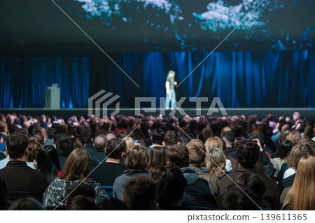 Large audience watching speaker on stage at technology conference with blue lighting and screen visuals Large audience watching speaker on stage at technology conference with blue lighting and screen visuals 139611365