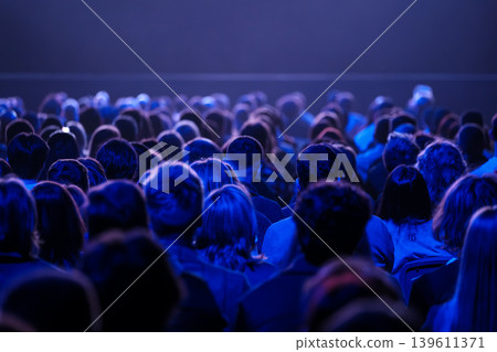 Audience watching stage under blue lighting, large seated crowd at concert or theater event Audience watching stage under blue lighting, large seated crowd at concert or theater event 139611371