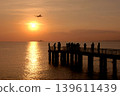 Sunset pier silhouettes with airplane landing over glowing horizon. Scenic evening view combining human presence, aviation, and serene water reflections in warm orange and pink tones. 139611439