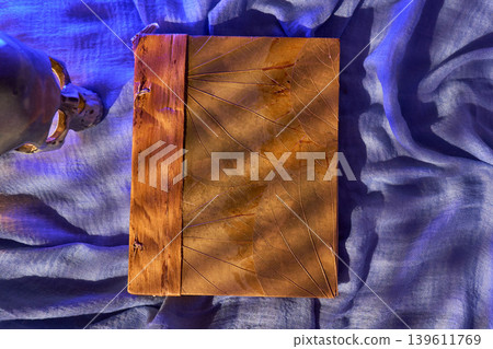 Sunlit ridges highlight handcrafted leather binding. Tactile details and handstitched spine on blue drape surface 139611769