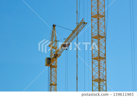 Two tall cranes stand in an urban area during the day. The cranes are positioned against a clear blue sky Two tall cranes stand in an urban area during the day. The cranes are positioned against a clear blue sky 139611965