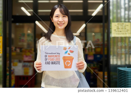 A young woman holding a sign about food waste in front of a supermarket. A young woman holding a sign about food waste in front of a supermarket. 139612401