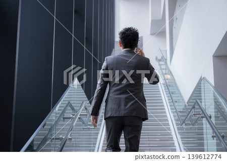 A middle-aged man with a beard, seen from behind, gives the impression of a businessman making a phone call while climbing the stairs in an office, showcasing his dynamism and speed in sales. A middle-aged man with a beard, seen from behind, gives the impression of a businessman making a phone call while climbing the stairs in an office, showcasing his dynamism and speed in sales. 139612774