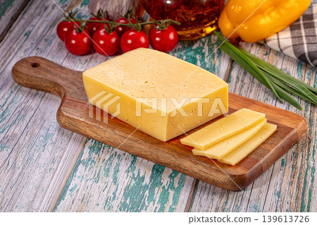 Sliced cheese on wooden cutting board with fresh vegetables and herbs on rustic table Sliced cheese on wooden cutting board with fresh vegetables and herbs on rustic table 139613726