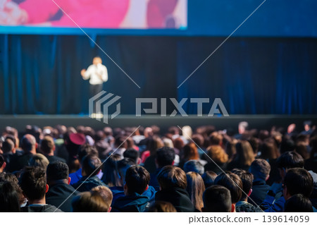 Audience watches speaker on stage at conference presentation in large dimly lit auditorium Audience watches speaker on stage at conference presentation in large dimly lit auditorium 139614059