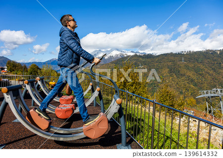 Man uses outdoor exercise machine on mountain with snow-capped peaks Man uses outdoor exercise machine on mountain with snow-capped peaks 139614332