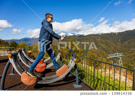 Man exercises on outdoor fitness equipment with a mountain view 139614336