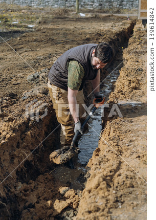 A man digs a drainage trench on his countryside property, working outdoors with a shovel 139614842
