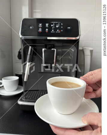 A pair of hands gently holds a white ceramic cup and saucer filled with freshly brewed coffee A pair of hands gently holds a white ceramic cup and saucer filled with freshly brewed coffee 139615126