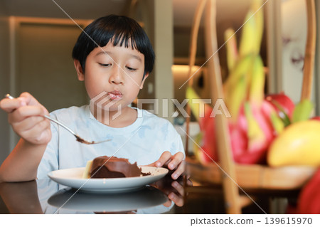 Cute Asian little boy happily eating delicious chocolate cake dessert with a fork at a dining table. Blurred tropical fruit basket in the foreground. Sweet childhood moments and enjoying tasty food. Cute Asian little boy happily eating delicious chocolate cake dessert with a fork at a dining table. Blurred tropical fruit basket in the foreground. Sweet childhood moments and enjoying tasty food. 139615970