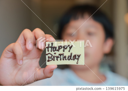 Close up child hand holding white chocolate piece with Happy Birthday sign written in dark chocolate. Blurred face of cute Asian boy in background. Sweet dessert for joyful childhood celebration. Close up child hand holding white chocolate piece with Happy Birthday sign written in dark chocolate. Blurred face of cute Asian boy in background. Sweet dessert for joyful childhood celebration. 139615975