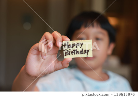 Close up child hand holding white chocolate piece with Happy Birthday sign written in dark chocolate. Blurred face of cute Asian boy in background. Sweet dessert for joyful childhood celebration. Close up child hand holding white chocolate piece with Happy Birthday sign written in dark chocolate. Blurred face of cute Asian boy in background. Sweet dessert for joyful childhood celebration. 139615976