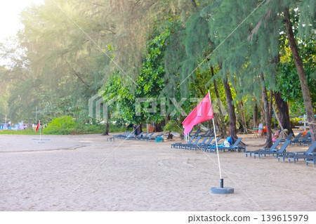 Empty tropical sandy beach resort with relaxing sun loungers under lush green pine trees shade and a red warning flag in foreground indicating no swimming on a bright sunny summer vacation holiday. Empty tropical sandy beach resort with relaxing sun loungers under lush green pine trees shade and a red warning flag in foreground indicating no swimming on a bright sunny summer vacation holiday. 139615979