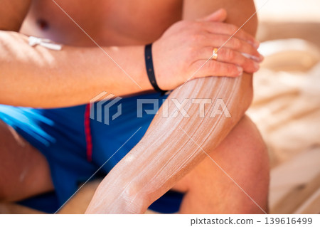 Caucasian man applying sunscreen cream on forearm while sitting on a beach during a summer vacation at a seaside resort. Sun protection and skincare routine 139616499