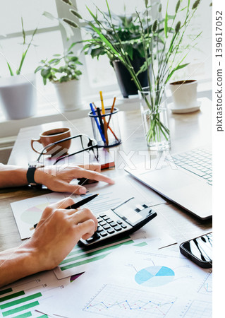 Financial analyst reviewing charts and using calculator at bright desk Financial analyst reviewing charts and using calculator at bright desk 139617052