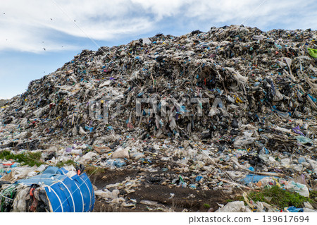 A landfill site shows towering mounds of waste with plastic and debris under a blue sky during daytime. Birds fly above the piles A landfill site shows towering mounds of waste with plastic and debris under a blue sky during daytime. Birds fly above the piles 139617694