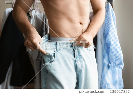 Man showing significant weight loss by pulling on his oversized jeans. Concept of body transformation, healthy lifestyle, and successful fitness progress. Man showing significant weight loss by pulling on his oversized jeans. Concept of body transformation, healthy lifestyle, and successful fitness progress. 139617832