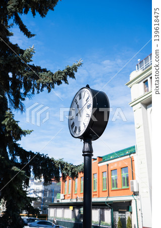 A classic black double-sided street clock with Roman numerals mounted on a metal pole. Vintage-style street clock in a city center against a blue sky. A classic black double-sided street clock with Roman numerals mounted on a metal pole. Vintage-style street clock in a city center against a blue sky. 139618475