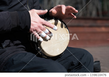 A close-up shot of a man's hands playing a traditional African djembe drum. The musician has white protective tape on his fingers. A close-up shot of a man's hands playing a traditional African djembe drum. The musician has white protective tape on his fingers. 139618476