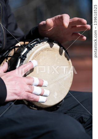 Close-up of a musician playing a djembe drum outdoors. Hands of a drummer with taped fingers playing an ethnic percussion instrument. Close-up of a musician playing a djembe drum outdoors. Hands of a drummer with taped fingers playing an ethnic percussion instrument. 139618525