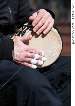 Close-up view of a musician playing a djembe drum outdoors. Hands of a drummer with taped fingers playing an ethnic percussion instrument. Close-up view of a musician playing a djembe drum outdoors. Hands of a drummer with taped fingers playing an ethnic percussion instrument. 139618529