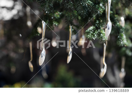 Surreal and whimsical garden scene with metallic spoons reflecting soft light. Multiple silver spoons hanging from evergreen tree branches in a garden. Silverware as unique tree ornaments. Surreal and whimsical garden scene with metallic spoons reflecting soft light. Multiple silver spoons hanging from evergreen tree branches in a garden. Silverware as unique tree ornaments. 139618889