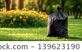 Garbage bag on a green lawn under sunlight surrounded by flowers 139623190