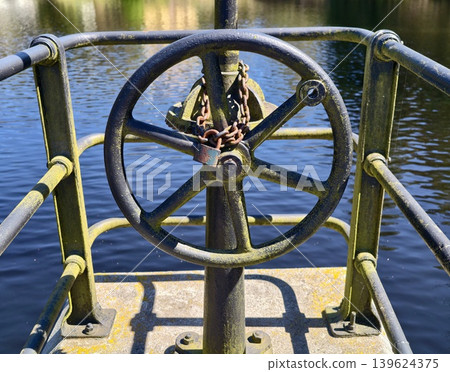 Aged Industrial Valve On Dockside Highlights Resilience Amid Tranquil Canal Setting And Weathered Surfaces Aged Industrial Valve On Dockside Highlights Resilience Amid Tranquil Canal Setting And Weathered Surfaces 139624375