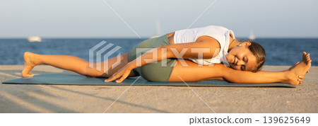 On windy day young slender woman does yoga near sea doing Hanumasana 139625649