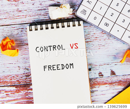 A spiral notepad shows control versus freedom in bold black and red letters. It rests on a rustic wooden table, next to a calculator and crumpled papers during the day 139625699