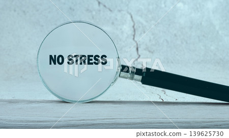 Observing the clear message No Stress through a magnifying glass placed on a clean wooden surface, set against a smooth light-colored wall Observing the clear message No Stress through a magnifying glass placed on a clean wooden surface, set against a smooth light-colored wall 139625730