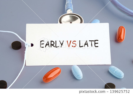 A white tag with the phrase Early versus Late highlights medical timing. Pills and a stethoscope rest on a gray background, emphasizing health choices A white tag with the phrase Early versus Late highlights medical timing. Pills and a stethoscope rest on a gray background, emphasizing health choices 139625752