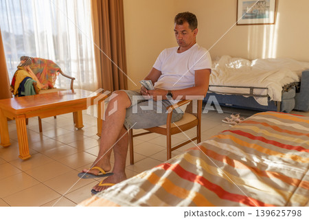 Portrait of middle-aged male tourist of Spanish appearance counts dollars while sitting on chair in hotel room 139625798