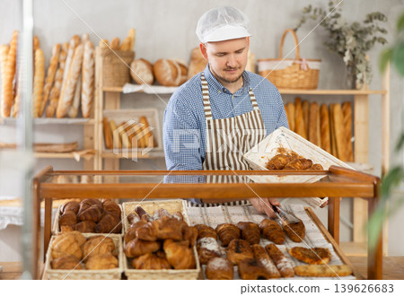 Man employee puts croissants in window, arranges display of goods at bakery. Man employee puts croissants in window, arranges display of goods at bakery. 139626683