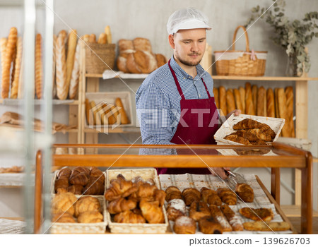 Man employee puts croissants in window, arranges display of goods at bakery. Man employee puts croissants in window, arranges display of goods at bakery. 139626703