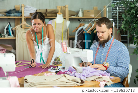 Male tailor sewing while young seamstress cuts textile Male tailor sewing while young seamstress cuts textile 139626718