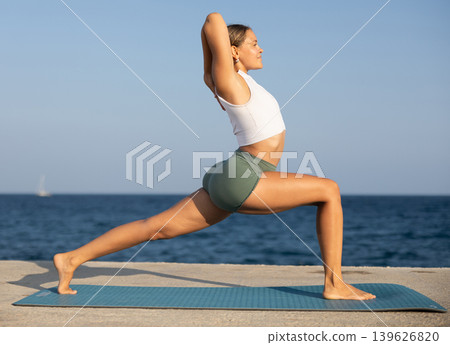 Girl practicing yoga on seashore, performing Anjaneyasana, or Crescent Moon Pose Girl practicing yoga on seashore, performing Anjaneyasana, or Crescent Moon Pose 139626820