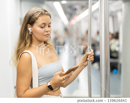 Young girl standing in subway wagon holding phone and wireless headphones on airs Young girl standing in subway wagon holding phone and wireless headphones on airs 139626841