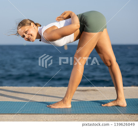 Sportive girl practicing forward bend pose of yoga on seashore Sportive girl practicing forward bend pose of yoga on seashore 139626849