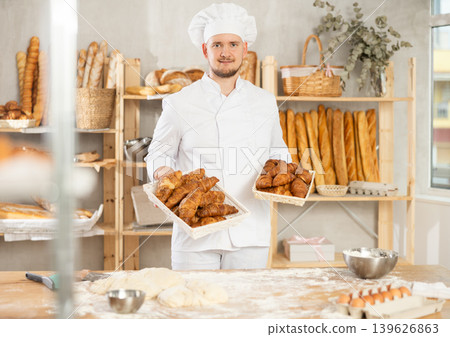 Baker in white uniform stands behind counter in small private bakery offering hot croissants Baker in white uniform stands behind counter in small private bakery offering hot croissants 139626863