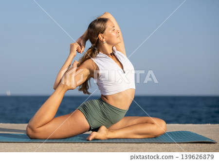 Sportive girl practicing king pigeon pose of yoga on seashore Sportive girl practicing king pigeon pose of yoga on seashore 139626879