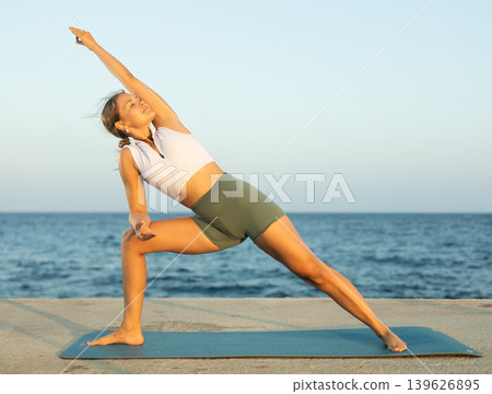 On windy day young slender woman does yoga near sea doing Utthita Parshvakonasana 139626895