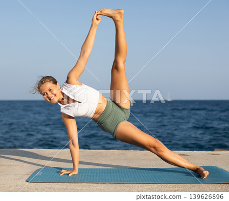 On windy day young slender woman does yoga near sea doing practicing hatha yoga 139626896