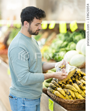 Customer stands near a fruit counter and chooses bananas in a supermarket Customer stands near a fruit counter and chooses bananas in a supermarket 139628954