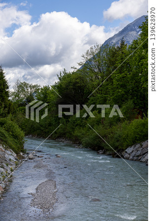 Alpine River Flowing Through Lush Green Forest Beneath a Cloudy Spring Sky 139628970