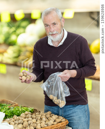 Elderly man stands at the counter of a supermarket and chooses walnuts. Elderly man stands at the counter of a supermarket and chooses walnuts. 139628986