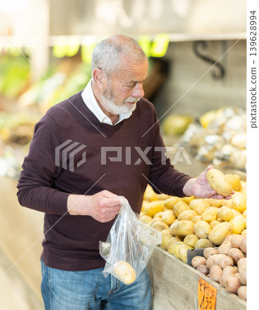 Elderly man visiting a vegetable store stands near the counter and chooses farm potato 139628994