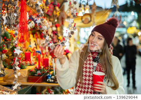 Girl with glass of coffee to go looks at wreaths and garlands of pine cones and red berries 139629044